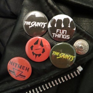 May include: Five round black and white buttons with different designs. The buttons have the text "The Saints", "Fun Things", "The Saints", and "Hitmen". One button has a black and white graphic of a symbol.