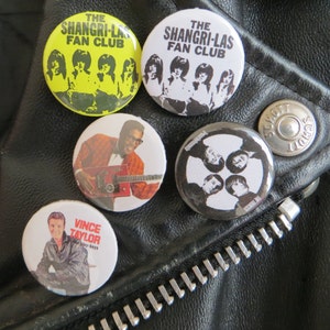 Könnte beinhalten: Vier gelbe und schwarze Anstecknadeln mit dem Text "The Shangri-Las Fan Club" und eine schwarz-weiße Anstecknadel mit dem Text "Vince Taylor - Art Boys".