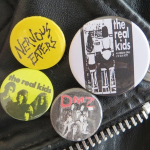May include: Four round pinback buttons with different designs. The top button is yellow with black text that reads "NERVOUS EATERS". The button to the right is white with black text that reads "the real kids ALL KINDA GIRLS  CW TAXI BOYS". The button to the left is yellow with black text that reads "the real kids". The bottom button is gray with red text that reads "DMZ".