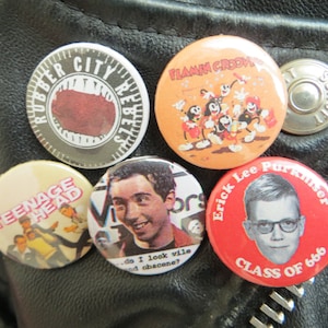 May include: Five round pinback buttons with various designs. The buttons are on a black leather jacket. The buttons feature a red mouth with the text "Rubber City Rebels", a cartoon scene with the text "Flamin Groovy", a black and white illustration of a person with the text "Teenage Head", a black and white illustration of a person with the text "do I look vile and obscene?", and a red button with the text "Erick Lee Purkiser Class of 666".