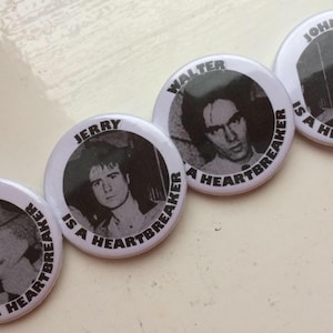 May include: Four white round buttons with black text and images of four different men. Each button says "[Name] is a Heartbreaker".