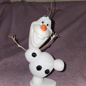 May include: A white snowman figurine with black eyes and buttons, an orange nose, and a wide smile. The snowman has twig arms and is standing on a white base.