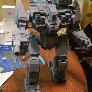 May include: A grey plastic model of a robot with a large gun on its left arm. The robot has a large, rectangular head with a small antenna on top. The robot is standing on a wooden table.