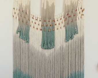 May include: Macrame wall hanging with a natural wood dowel. The design features a gradient of colors, transitioning from white to gray and ending in a teal fringe. Wooden beads are woven into the upper section.