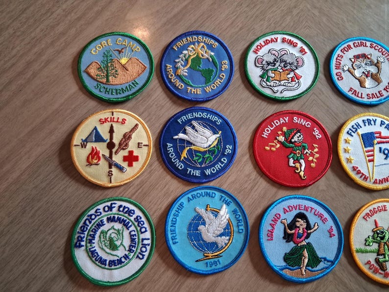 Pu&ograve; includere: Una collezione di toppe ricamate colorate con vari disegni. Alcune toppe presentano testi come "Gore Camp Scherman", "Friendships Around the World" e "Holiday Sing '92". Altre raffigurano immagini di animali e scene.