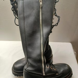 May include: A pair of black leather knee-high boots with silver zippers and laces. The boots have a round toe and a low heel. The zippers run down the front of the boots. The laces are on the front of the boots.