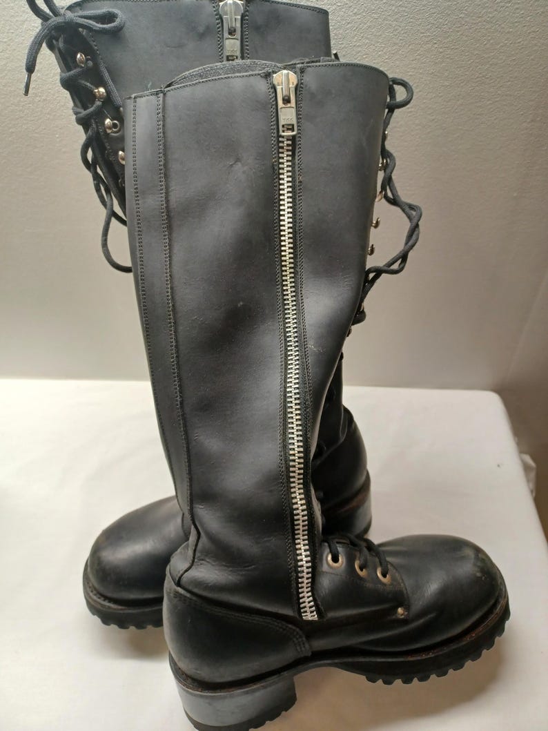 May include: A pair of black leather knee-high boots with silver zippers and laces. The boots have a round toe and a low heel. The zippers run down the front of the boots. The laces are on the front of the boots.
