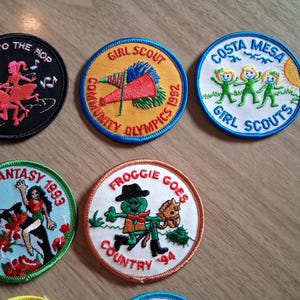 Pu&ograve; includere: Una collezione di toppe ricamate con vari disegni e testo. Una toppa dice "Let's Go to the Hop" con figure danzanti. Altre toppe includono "Girl Scout", "Costa Mesa Girl Scouts", "High Seas Fantasy 1993" e "Froggie Goes Country '94".