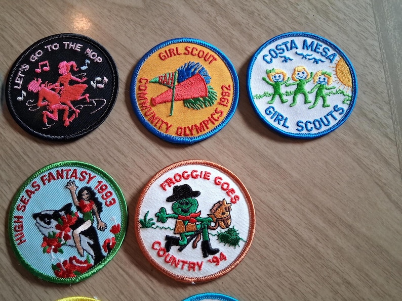 Pu&ograve; includere: Una collezione di toppe ricamate con vari disegni e testo. Una toppa dice "Let's Go to the Hop" con figure danzanti. Altre toppe includono "Girl Scout", "Costa Mesa Girl Scouts", "High Seas Fantasy 1993" e "Froggie Goes Country '94".