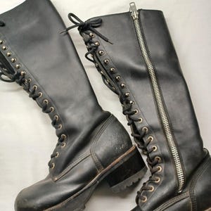 May include: Pair of black leather knee-high boots. The boots feature a lace-up design with metal eyelets, a side zipper, and a chunky lug sole. The boots have a worn appearance.