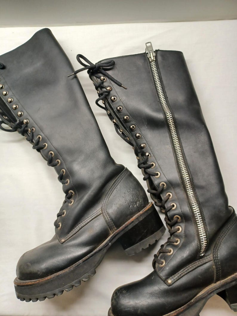 May include: Pair of black leather knee-high boots. The boots feature a lace-up design with metal eyelets, a side zipper, and a chunky lug sole. The boots have a worn appearance.