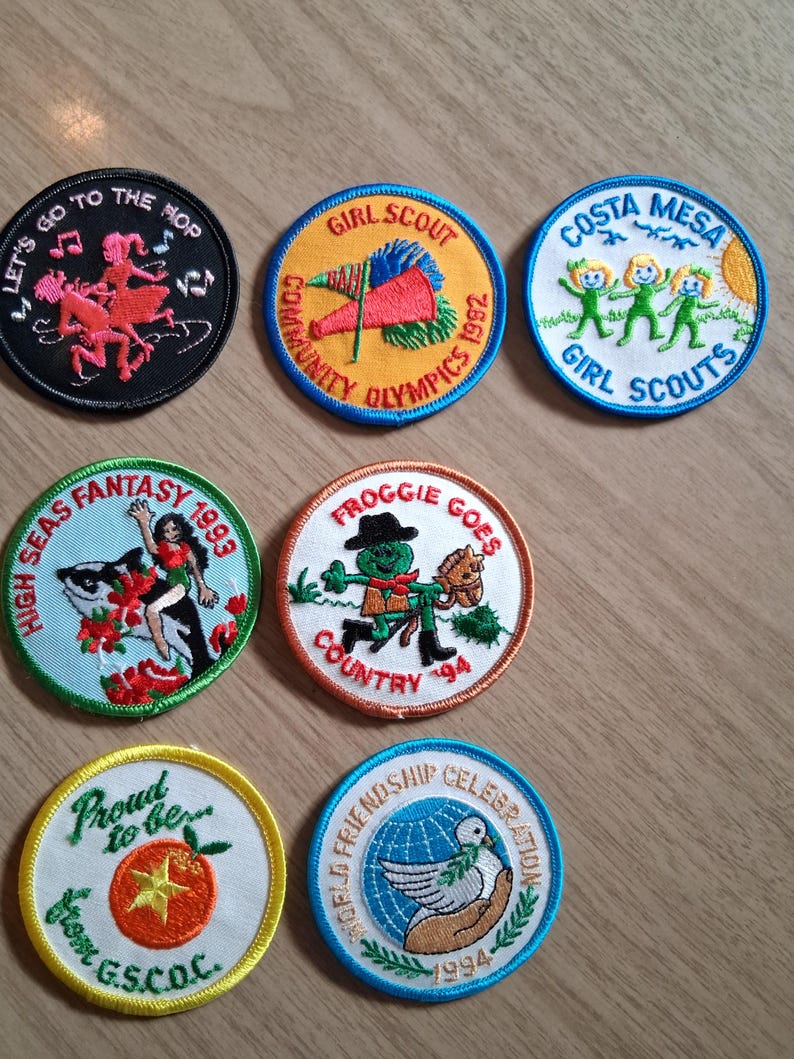 Pu&ograve; includere: Una collezione di toppe colorate delle Girl Scout. I disegni includono una figura danzante, una bandiera, tre figure verdi, una donna con una balena, una rana, un pomodoro e una colomba. Testi: "Let's Go To The Hop", "Girl Scout", "Costa Mesa Girl Scouts", "High Seas Fantasy", "Froggie Goes Country".