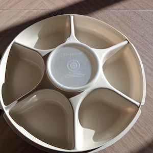 May include: A beige plastic serving tray with a central circular compartment and five surrounding sections. The center has a lid with the Tupperware logo. The tray is designed for serving snacks or appetizers.