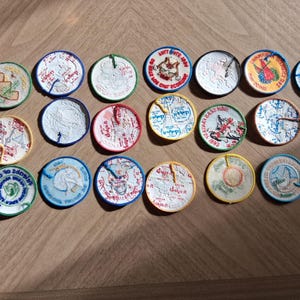 Pu&ograve; includere: Una collezione di ventuno toppe rotonde ricamate con vari disegni e colori. Le toppe presentano immagini di animali, testo e altre grafiche. Le toppe sono disposte su una superficie di legno.