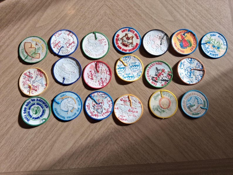 Pu&ograve; includere: Una collezione di ventuno toppe rotonde ricamate con vari disegni e colori. Le toppe presentano immagini di animali, testo e altre grafiche. Le toppe sono disposte su una superficie di legno.