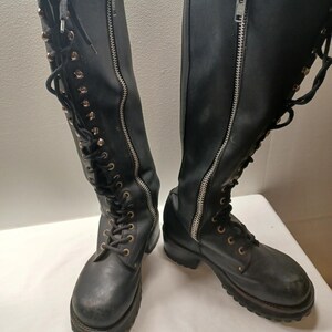 May include: A pair of black knee-high boots with silver zippers and laces. The boots have a rugged, worn appearance, with visible stitching and metal eyelets. The soles are thick and textured.