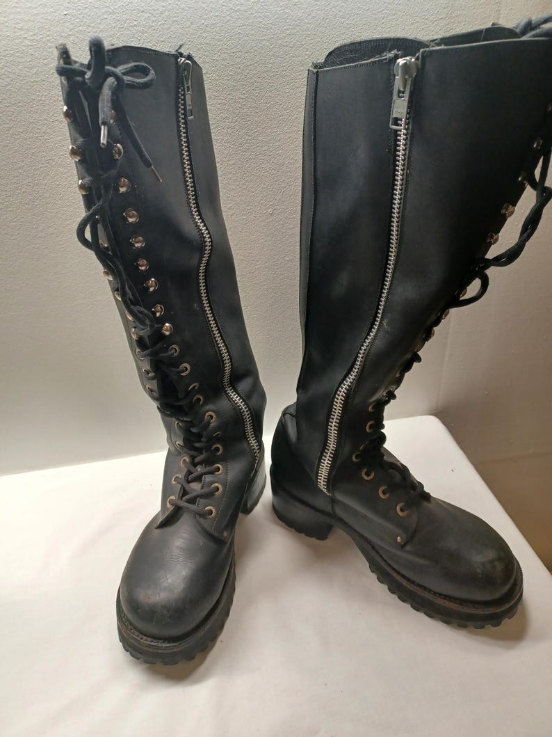 May include: A pair of black knee-high boots with silver zippers and laces. The boots have a rugged, worn appearance, with visible stitching and metal eyelets. The soles are thick and textured.