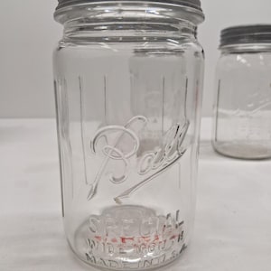Pode incluir: Frasco de vidro transparente Ball com tampa de metal. O frasco tem a palavra "Ball" em relevo na frente, juntamente com "SPECIAL WIDE MOUTH MADE IN U.S.A." O frasco é projetado para preservar alimentos.