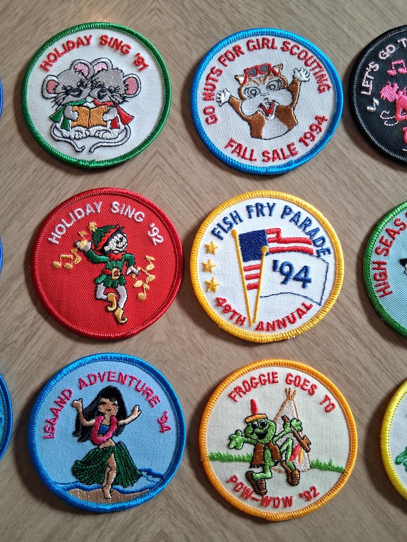 Pu&ograve; includere: Una collezione di toppe ricamate con vari disegni e testo. Le toppe presentano immagini di animali, persone e testo come "Holiday Sing", "Go Nuts for Girl Scouting" e "Island Adventure". Le toppe sono circolari con bordi colorati.