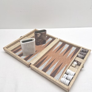 May include: Open backgammon board with a tan, brown, and silver triangular pattern. Includes dice, dice cups, and game pieces. The board is open, revealing the playing surface and storage compartments.