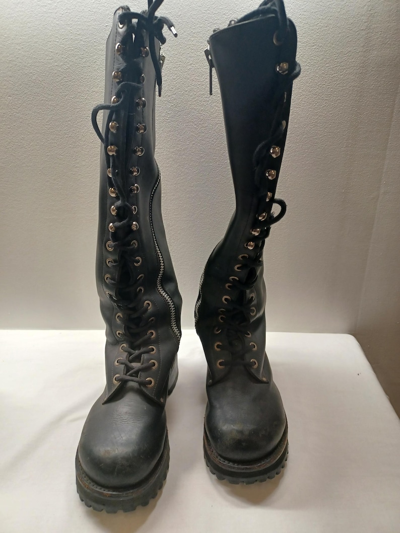May include: A pair of black knee-high boots with a lace-up design and side zippers. The boots have a rugged sole and metal eyelets. The laces are black, and the zippers are silver.