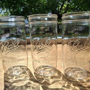 May include: Three clear glass Ball wide mouth mason jars arranged in a row. Each jar features the embossed Ball logo on the front, along with measurement markings in ounces and cups. The jars have a wide opening and are made in the USA.