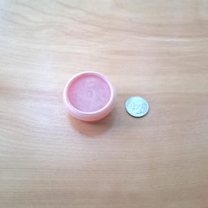 May include: A small, round, light pink plastic container filled with a translucent pink substance, positioned next to a silver-colored United States quarter coin for scale, showing the relative size of the circular cosmetic or balm jar.