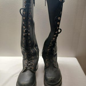May include: A pair of black knee-high boots with a lace-up design and side zippers. The boots have a rugged sole and metal eyelets. The laces are black, and the zippers are silver.