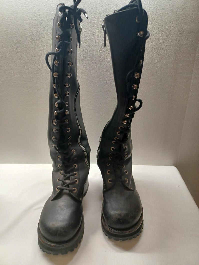May include: A pair of black knee-high boots with a lace-up design and side zippers. The boots have a rugged sole and metal eyelets. The laces are black, and the zippers are silver.