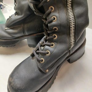 May include: Black leather boots with a lace-up front and a side zipper. The boots have a chunky sole and metal eyelets. The boots appear worn, with scuffs and marks on the leather.