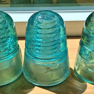 Op de afbeelding: Drie vintage aquablauwe glazen elektrische isolatoren met een afgeronde koepelvormige bovenkant en een schroefdraad aan de binnenkant. Elk stuk heeft een geribbeld exterieurontwerp en reliëfletters aan de zijkant, met een doorschijnende afwerking en een solide constructie.