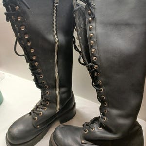 May include: Pair of black leather knee-high boots with silver-tone grommets and laces. One boot features a full-length zipper. The boots have a chunky sole and a slight heel, suggesting a rugged, alternative style.