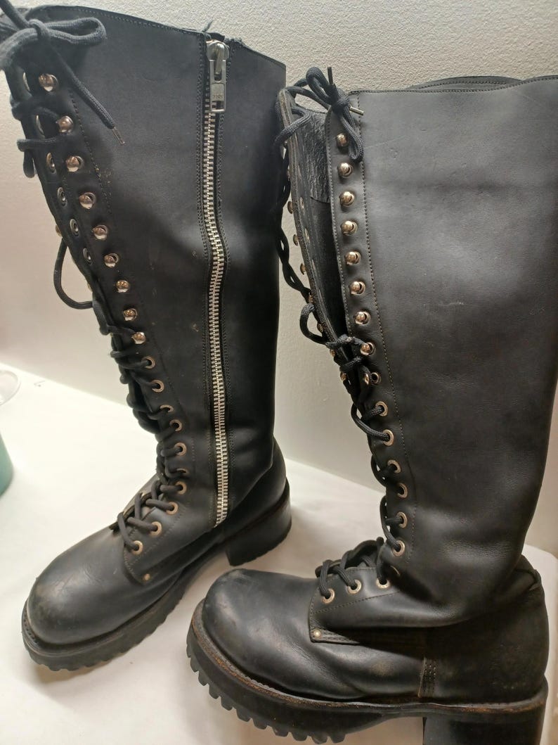 May include: Pair of black leather knee-high boots with silver-tone grommets and laces. One boot features a full-length zipper. The boots have a chunky sole and a slight heel, suggesting a rugged, alternative style.