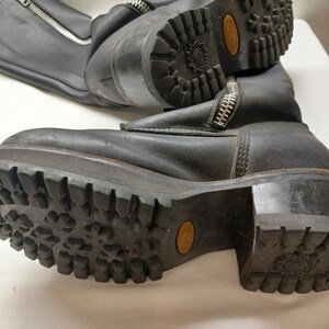 May include: Pair of black leather boots with chunky soles and side zippers. The boots have a rugged, worn appearance, with visible wear on the soles and uppers. The soles have a deep tread pattern for traction. The boots are likely for outdoor or work wear.