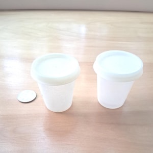 May include: Two small, translucent white plastic cups with matching opaque white lids, positioned next to a single silver-colored coin for scale. The cups feature a tapered cylindrical shape and a smooth, simple design for storage or small portions.