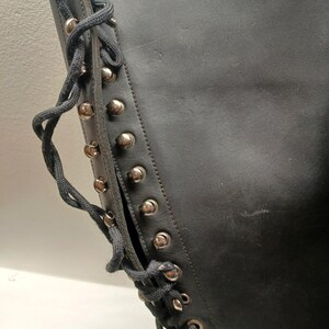 May include: Close-up of a black leather boot with silver-colored studs and dark gray laces. The boot has a side opening and a high shaft. The laces are threaded through metal eyelets.