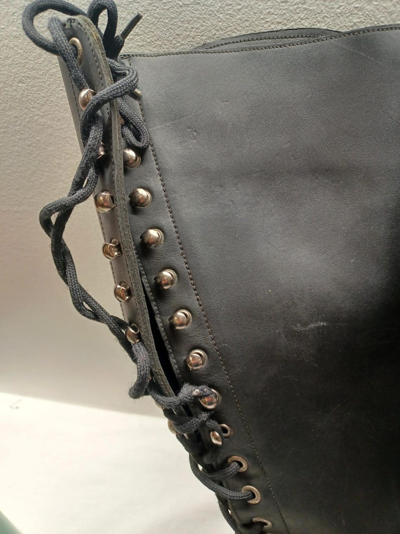 May include: Close-up of a black leather boot with silver-colored studs and dark gray laces. The boot has a side opening and a high shaft. The laces are threaded through metal eyelets.
