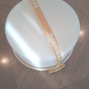 Puede incluir: Un portatartas redondo de color crema con un asa dorada. El portatartas tiene una tapa y una base, y el asa está adornada con círculos decorativos. El diseño es sencillo y funcional.