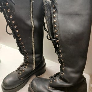 May include: A pair of black leather knee-high boots. The boots feature a lace-up design with metal eyelets and a side zipper. The soles are thick and lugged. The boots have a classic, rugged style.