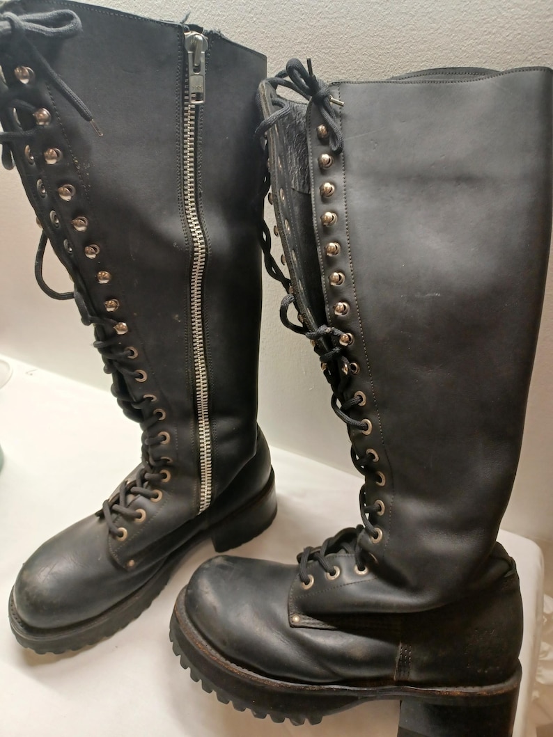 May include: A pair of black leather knee-high boots. The boots feature a lace-up design with metal eyelets and a side zipper. The soles are thick and lugged. The boots have a classic, rugged style.