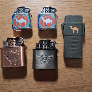 May include: A collection of five metal lighters with various designs. Two lighters have a camel logo on a blue and orange background. Other lighters feature a copper finish, a silver finish with wings, and an olive green case with a camel logo.