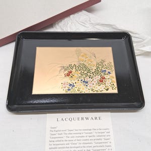 May include: A rectangular black lacquerware tray featuring a central gold panel decorated with a colorful floral and grass motif. The tray is accompanied by a white informational card with the text LACQUERWARE and a description of the term.