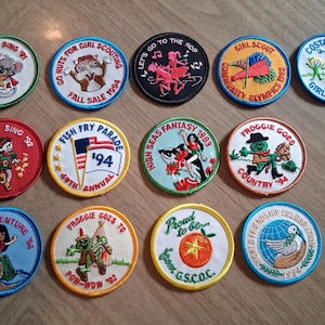 Pu&ograve; includere: Una collezione di toppe ricamate con vari disegni e testo. Le toppe presentano immagini di animali, bandiere e personaggi dei cartoni animati. Il testo include "Holiday Sing '91", "Fish Fry Parade '94" e "Girl Scout Community Olympics 1992".