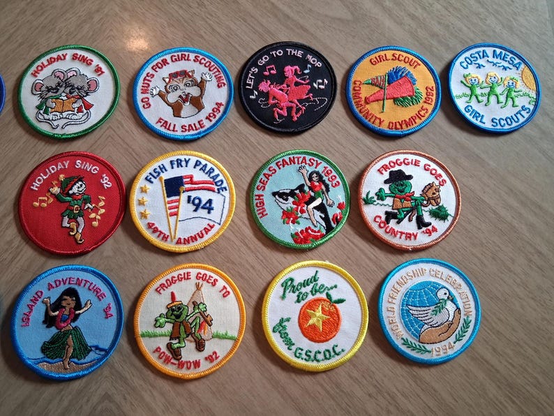Pu&ograve; includere: Una collezione di toppe ricamate con vari disegni e testo. Le toppe presentano immagini di animali, bandiere e personaggi dei cartoni animati. Il testo include "Holiday Sing '91", "Fish Fry Parade '94" e "Girl Scout Community Olympics 1992".