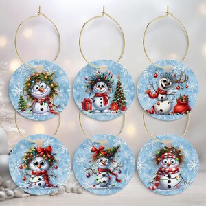May include: Set of six Christmas wine glass charms. Each charm features a round, light blue design with a snowman illustration, snowflakes, and a gold-colored hoop. The snowmen have festive accessories like scarves and lights.