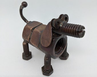 May include: A bronze-coloured metal dog sculpture, crafted from nuts, bolts, and screws. The sculpture has a screw snout, bolt legs, and a coiled tail. The dog is made from metal and stands on a white surface.