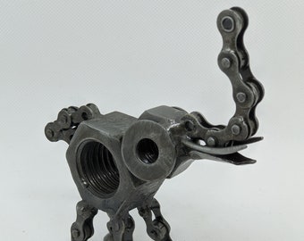 May include: A handcrafted metal elephant sculpture. The body is a large hex nut, with a smaller nut forming the head. Bicycle chain links create the trunk, legs, and ears. The sculpture is a unique piece of industrial art.