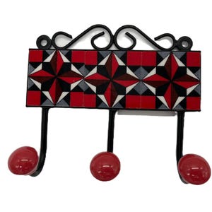 May include: A black metal wall hook with three hooks and red ceramic knobs. The backplate features a red, black and white geometric tile pattern.