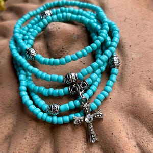 May include: A multi-strand bracelet made of small, round, bright turquoise beads. The bracelet features several silver-toned metal spacer beads with intricate patterns and a central silver-toned cross pendant adorned with small, clear, sparkling crystals.
