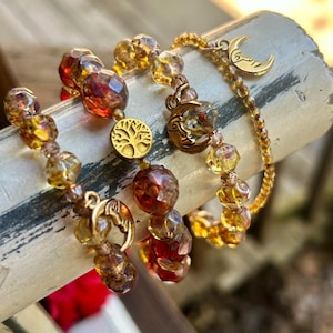 May include: A set of three beaded bracelets featuring faceted amber, orange, and red glass beads. The bracelets include gold-toned metal charms, specifically a circular tree of life emblem and two crescent moon charms with engraved facial features.
